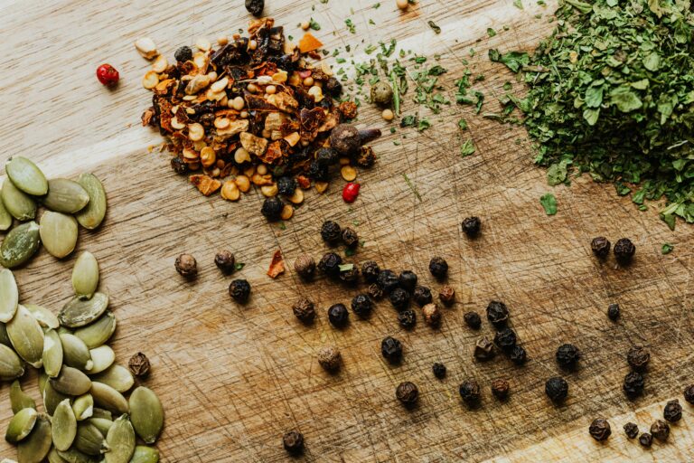 A close-up of various herbs and spices including peppercorns and pumpkin seeds on a rustic wooden surface.